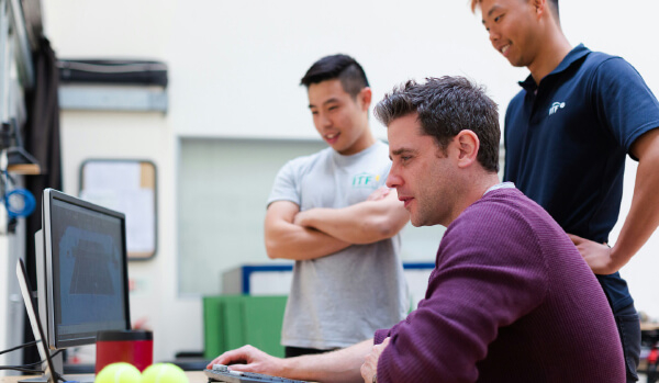 Three men are gathered around a computer, with one man sitting and focused on the screen while the other two stand nearby, observing.