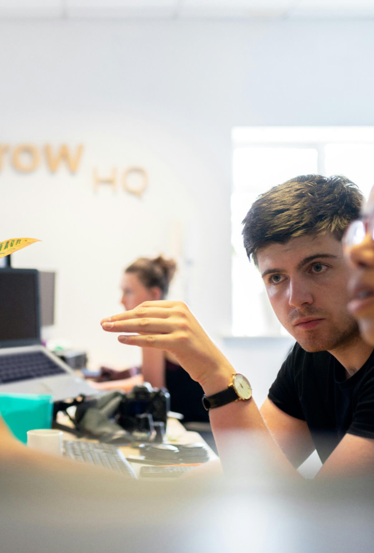 Two people sit at a desk having a discussion. Another person works at a computer in the background.