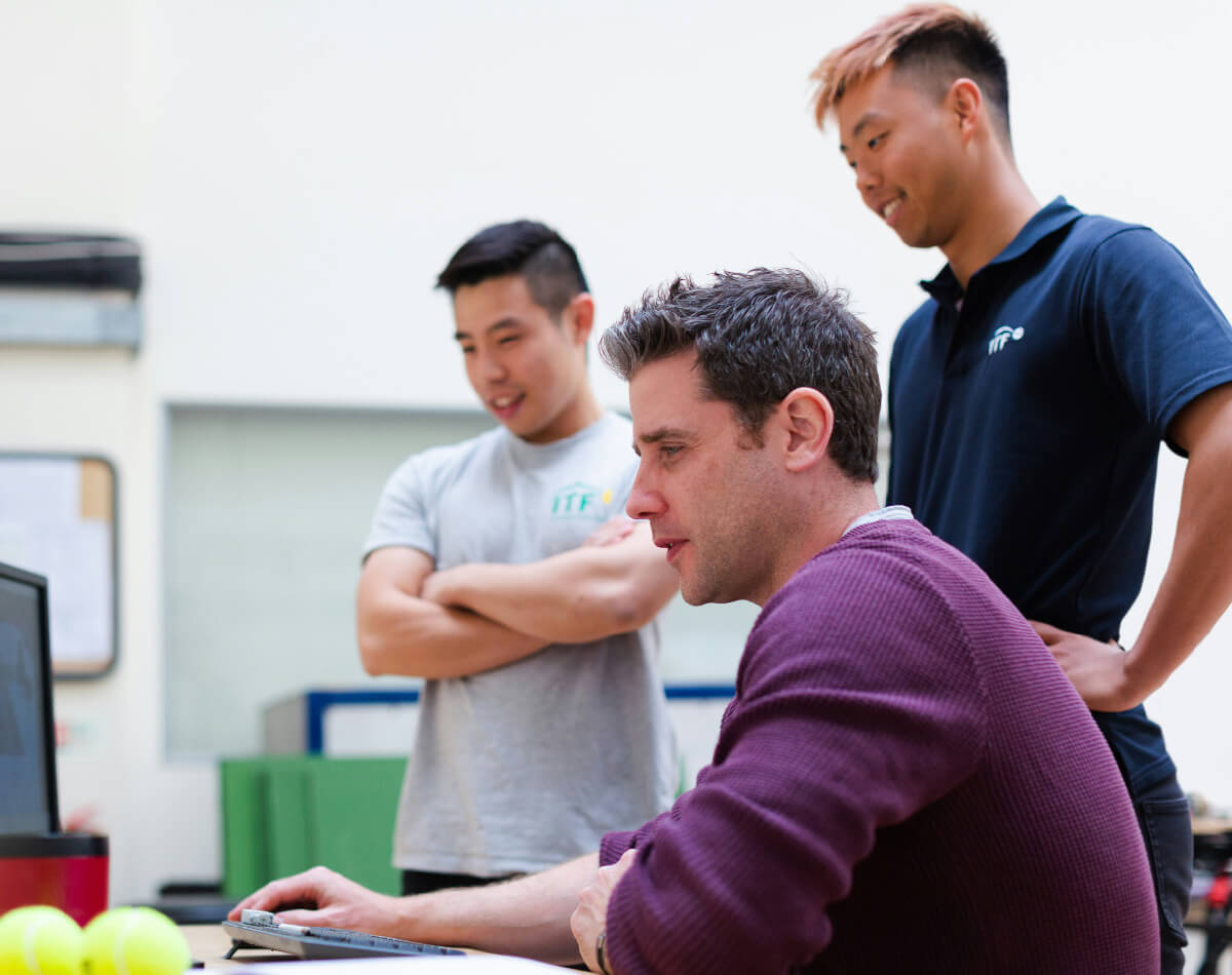 Three men are gathered around a computer, with one man sitting and focused on the screen while the other two stand nearby, observing.