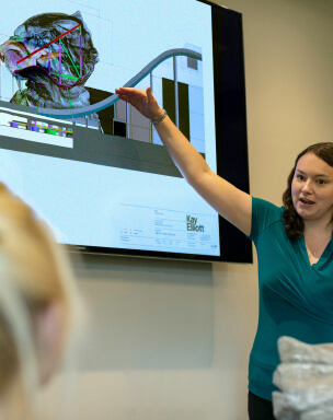 A woman gestures while presenting an image on a screen that appears to depict an architectural design.