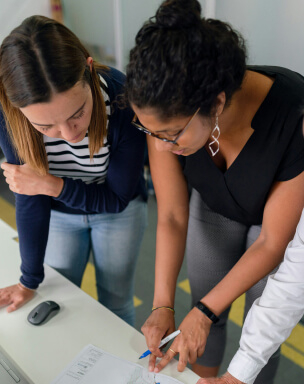 Two women and a man are gathered around a desk, examining a document and discussing it, with one woman pointing at the paper.