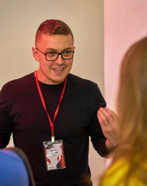 A man with short hair and glasses wearing a black shirt and a red lanyard speaks animatedly to someone off-frame.