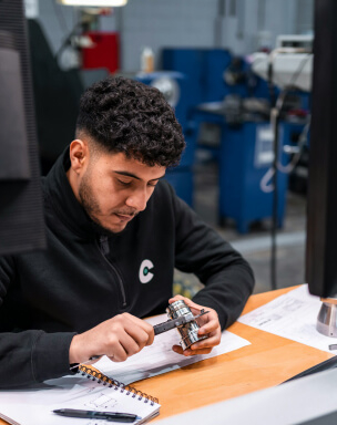 A young man is using a caliper to measure a mechanical part while sitting at a desk in a workshop, with a notebook and a pencil nearby.