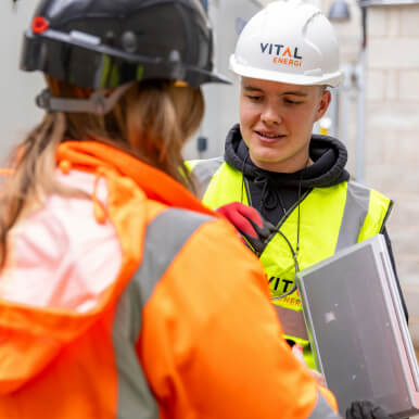 Two construction workers in safety gear, one wearing a helmet and high-visibility vest, interacting on a job site.