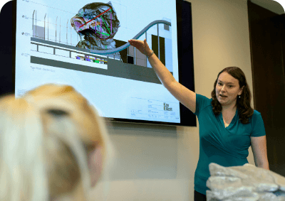 A woman gestures towards a presentation on a screen displaying a complex graphic, while another person sits looking on..
