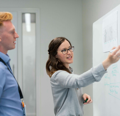 A woman writes on a whiteboard while talking to a man in a professional office setting.