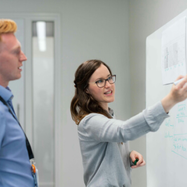 A woman writes on a whiteboard while talking to a man in a professional office setting.
