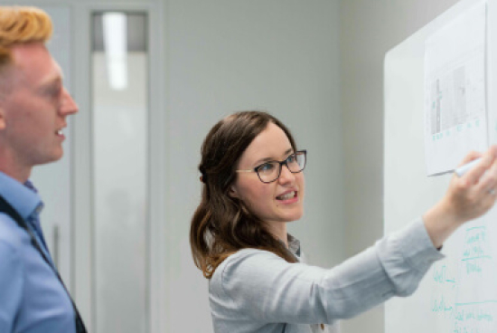 Two people engaging with work on a whiteboard. One is pointing to the board.