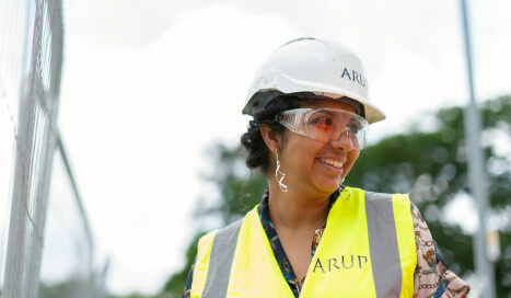 A woman wearing a safety helmet and goggles, with a yellow high-visibility vest, smiling at a construction site.