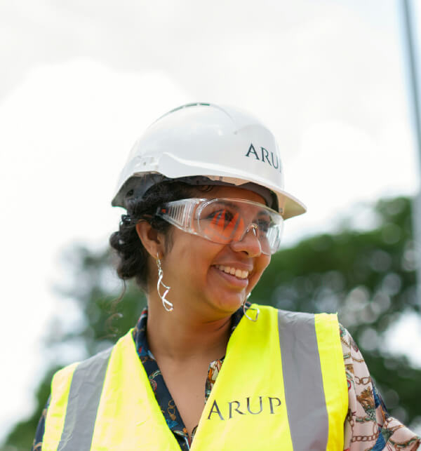 A woman wearing a safety helmet and glasses, smiling while dressed in a high-visibility vest.