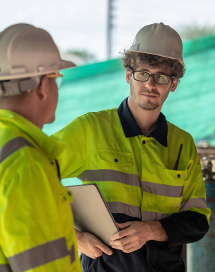 Two construction workers in safety gear, including hard hats and high-visibility jackets, discussing at a construction site. One worker is holding a tablet.