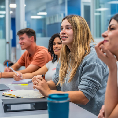 A diverse group of students engaged in a classroom setting, sitting at a table with notebooks and a laptop, displaying expressions of interest and participation.