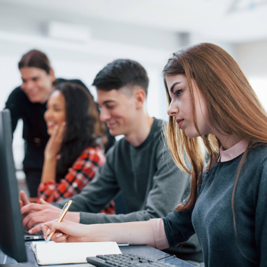 A group of students working together in a classroom, focused on computer screens, with one student taking notes.