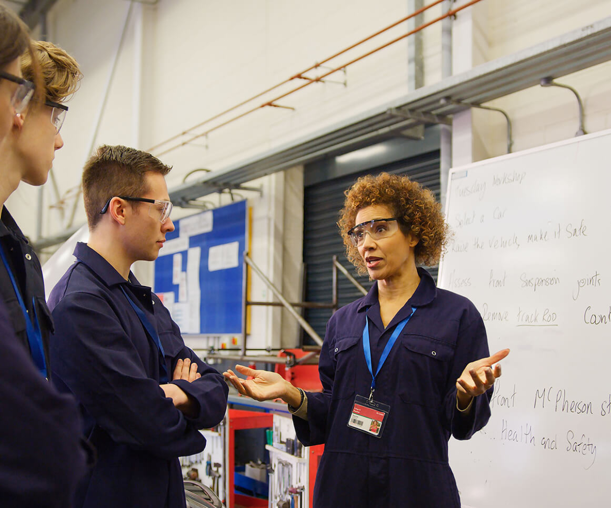 A group of students in blue work overalls listening to an instructor in a workshop setting, discussing a technical topic.