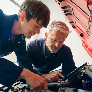 Two mechanics, a younger man and an older man, are working together under the hood of a car, using tools and looking at a tablet.