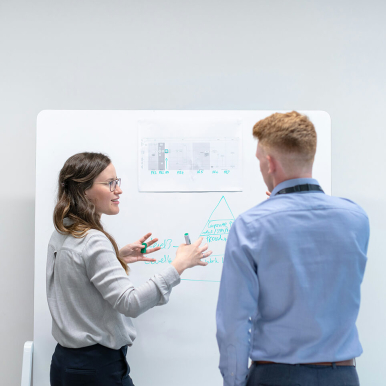 Two people are standing in front of a whiteboard, discussing a chart or diagram. One person is gesturing while pointing at the board, and the other is listening attentively.