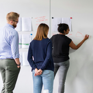 Three individuals are standing in front of a whiteboard filled with sketches and notes. One person is drawing on the whiteboard, while the others observe.