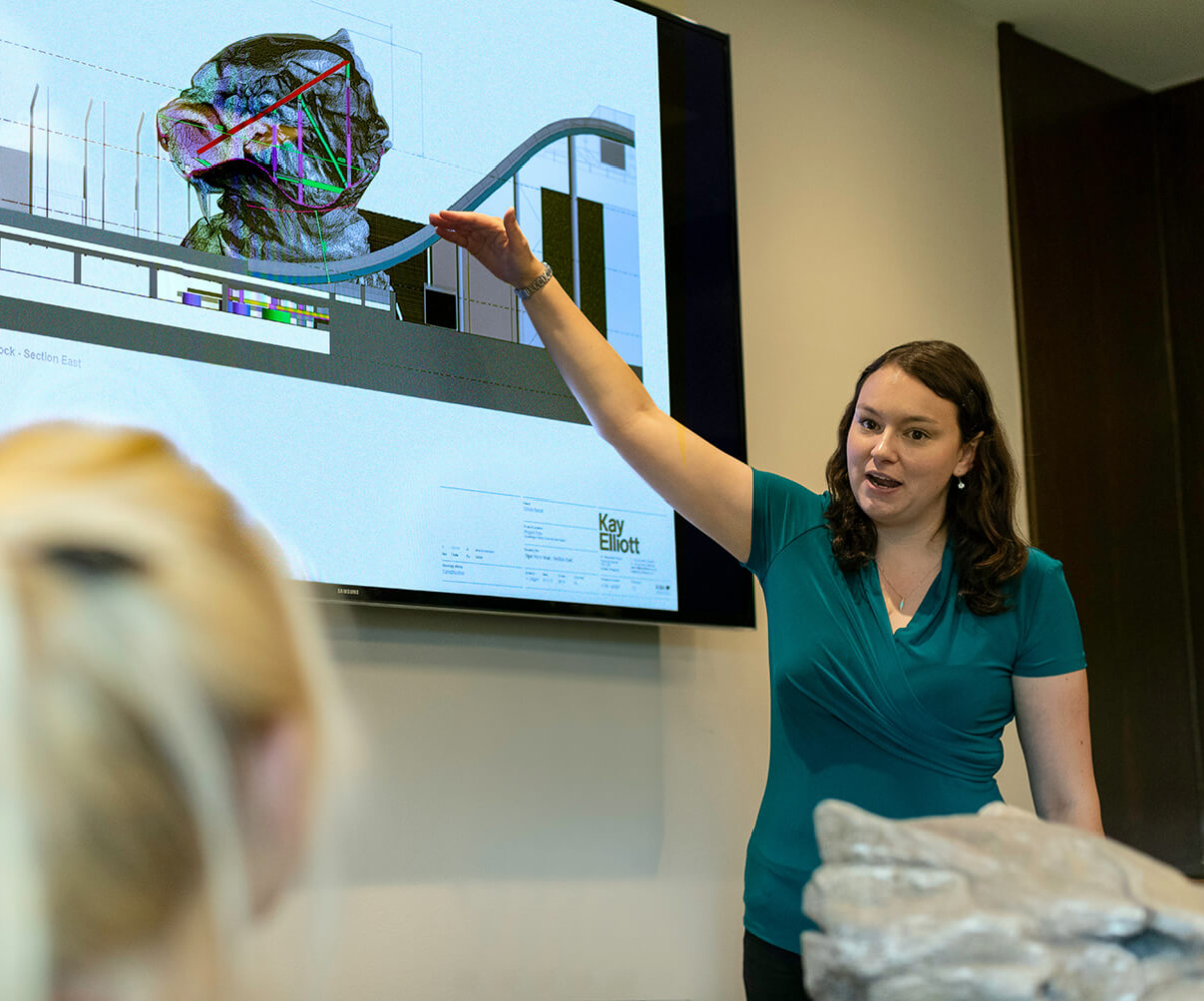 A woman presenting in front of a screen displaying architectural graphics, with her hand raised to emphasise a point.
