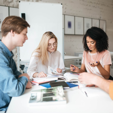 Four young adults engaged in a collaborative study session, reviewing books and notes at a table in a bright, modern workspace.