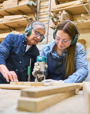A teacher and a student working together in a woodworking shop, using tools and focusing on a project. The student is using a rotary tool, while the teacher is observing and offering guidance.