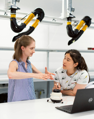 Two women in a laboratory setting, one demonstrating a device while the other observes, with equipment and a laptop on the table.
