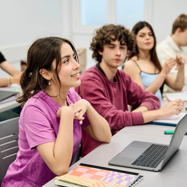A group of students sitting in a classroom, with one girl in the foreground smiling and engaging with someone out of frame..