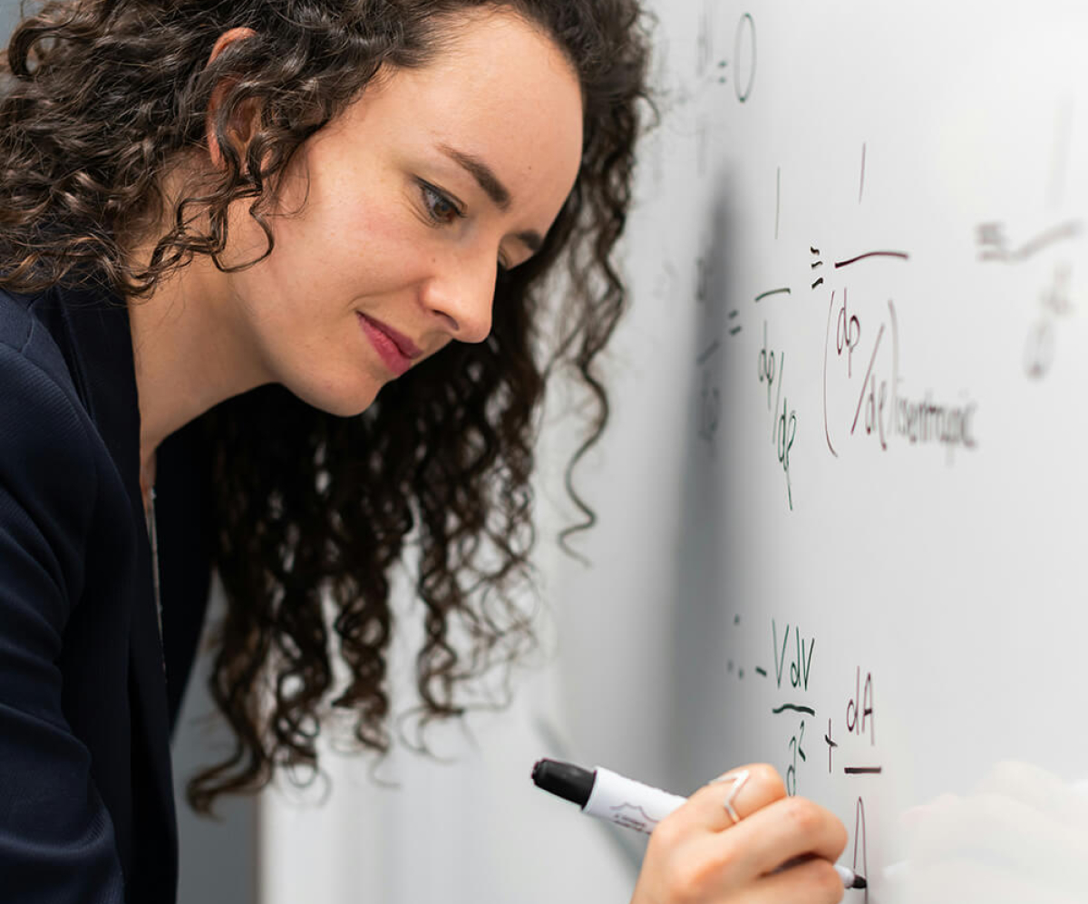 A woman wearing a dark blazer writes mathematical equations on a whiteboard using a marker.