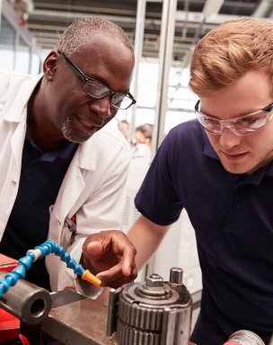 Two men in a workshop, one older and one younger, working on a machine with tools. The older man is guiding the younger one, both wearing safety glasses and focused on their task.