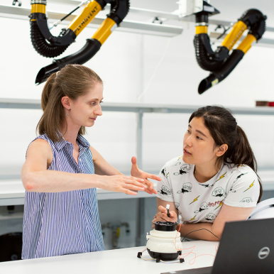 Two women in a lab setting, one gesturing while explaining something, and the other listening intently.