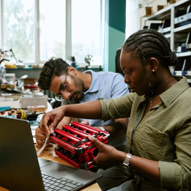 Two people sit in front of a laptop whilst modifying a machine placed on the desk in front of them.